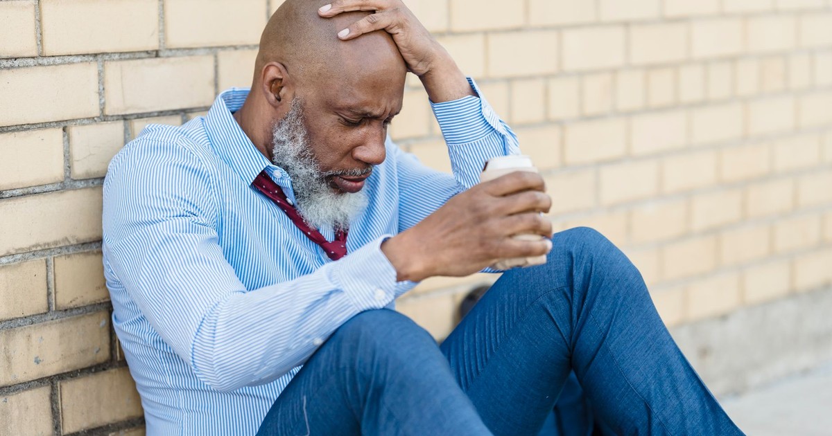 An elderly man in a suit sitting against a brick wall with a drink, appearing stressed.
