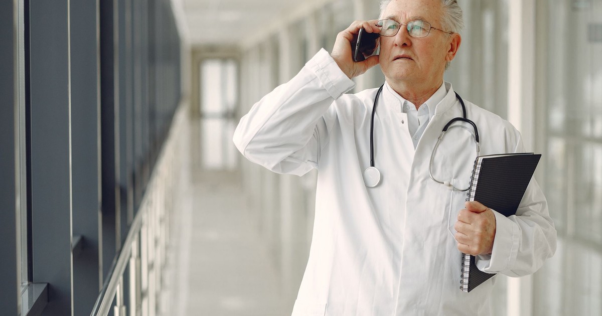 Serious mature male doctor in medical uniform standing with stethoscope and notebook while talking on smartphone in clinic corridor