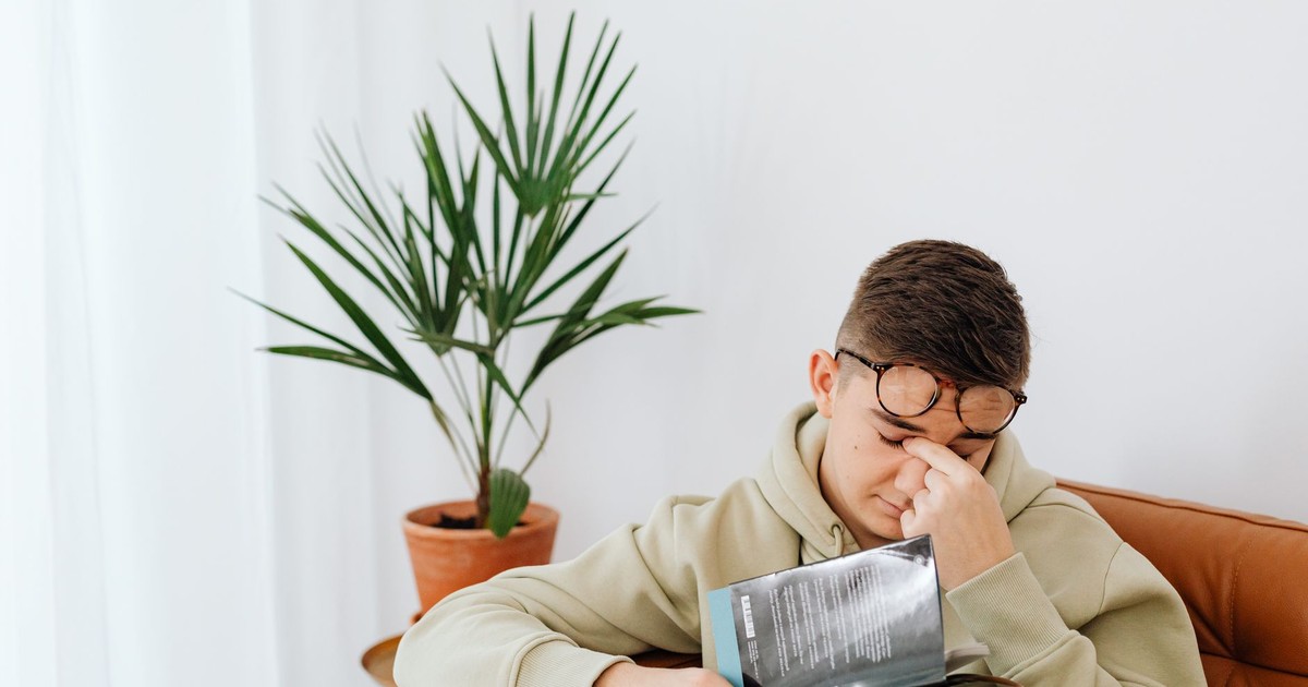Adult man wearing hoodie rubs his eyes while reading a book indoors, next to a potted plant.