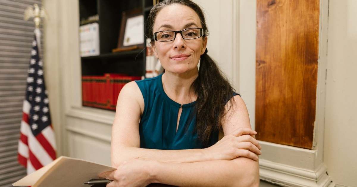Woman in a blue top with glasses smiling in an office with an American flag.