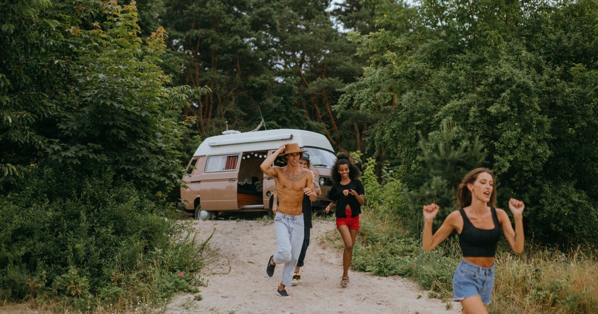 Energetic friends running near a camper van in a lush forest, enjoying a summer adventure.