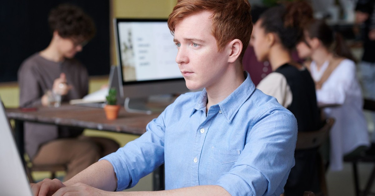 Young professional working intently on a computer in a busy modern office setting, surrounded by colleagues.