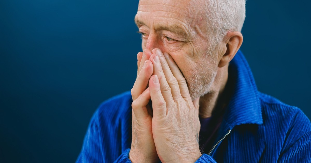 Serious anxious bearded senior gray haired male in blue jacket covering mouth with hands while looking away against blue background