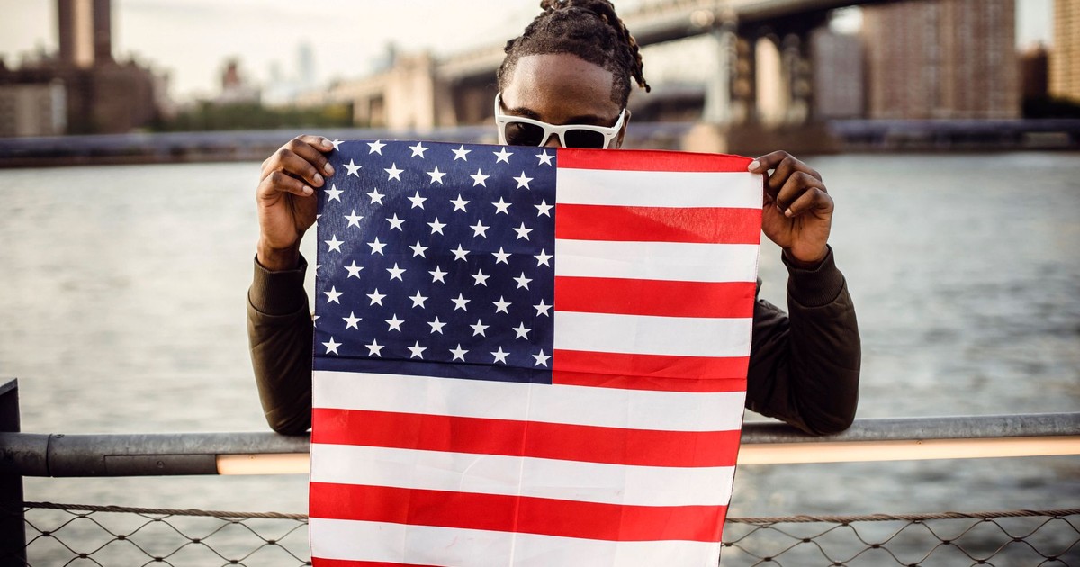 Ethnic male in casual clothes and sunglasses standing on embankment of city river while leaning on fence showing national flag of United States of America
