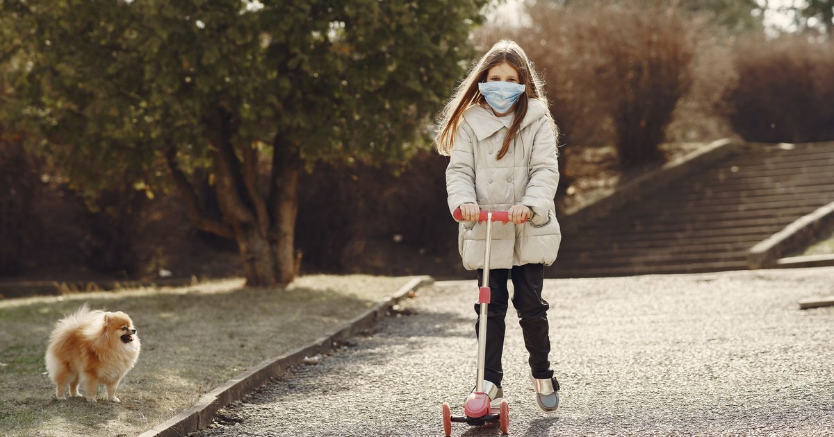 Full body of kid in warm wear and protective mask riding scooter near adorable Pomeranian dog looking at camera in park