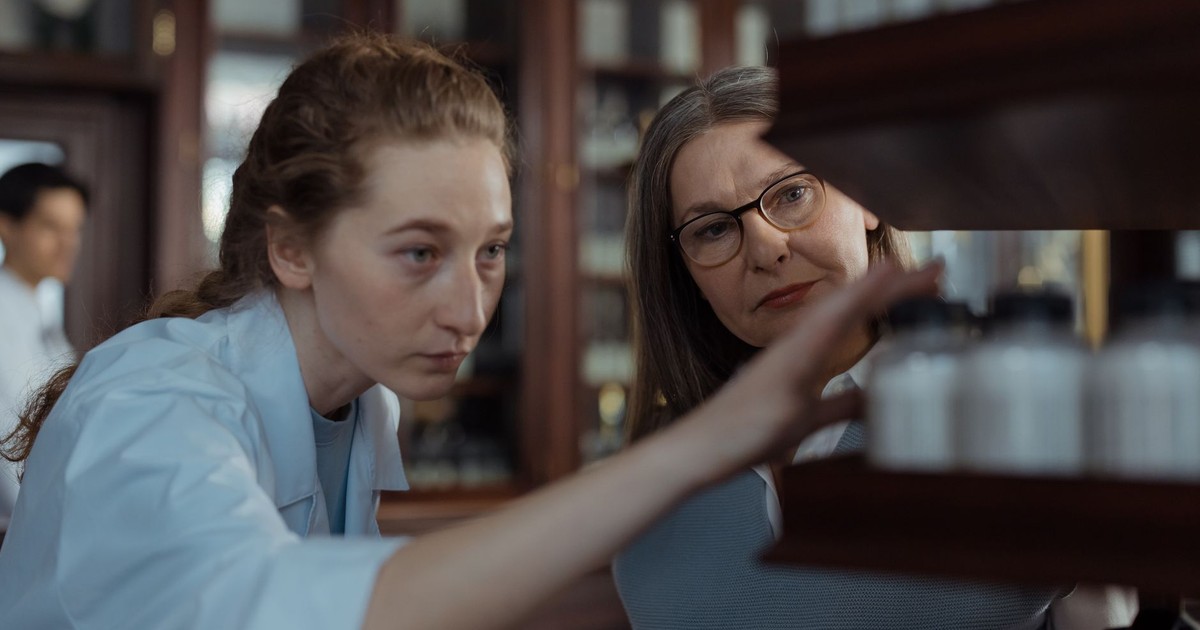 Two pharmacists wearing white coats and glasses examining medicine bottles on a pharmacy shelf.