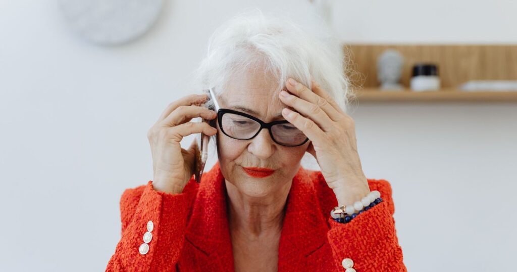 Senior woman in red blazer stressed while talking on the phone indoors.