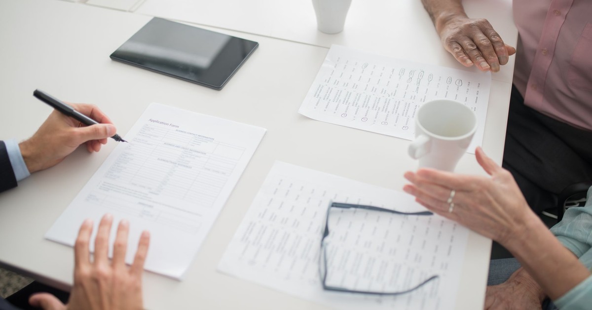 Hands writing on documents in a business meeting setup with coffee cups and tablet.