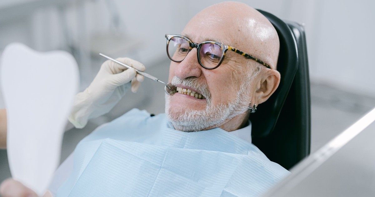 Elderly man at dental appointment, examining teeth with mirror.