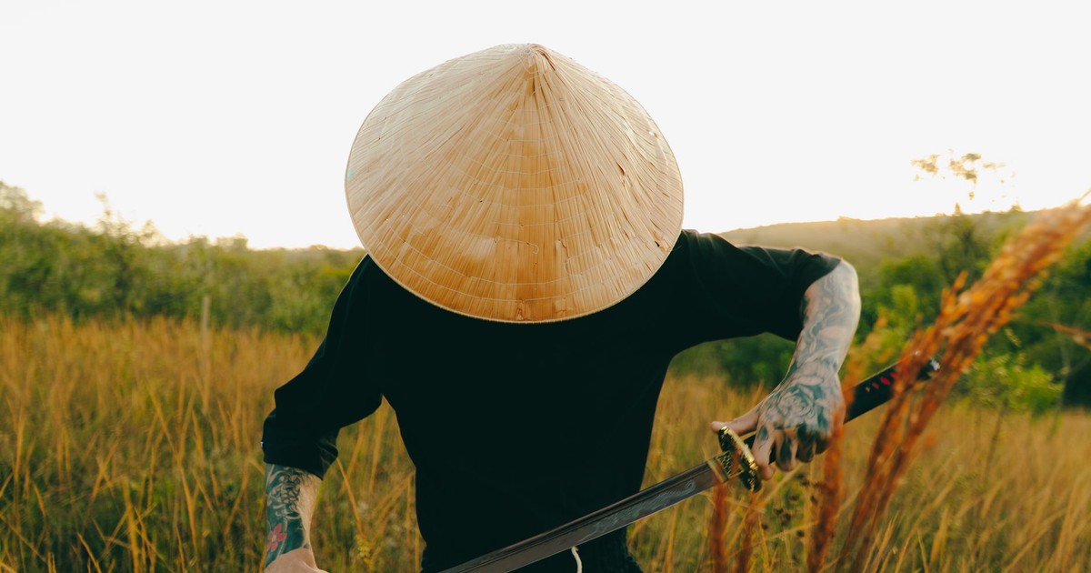 A tattooed individual wearing a conical hat with a samurai sword in a grassy outdoor setting.