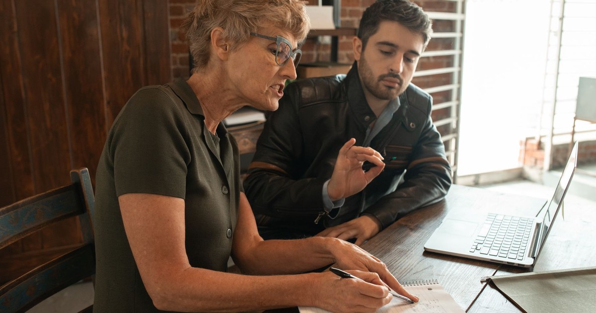 Senior woman guiding adult man during an indoor mentorship session with a laptop.