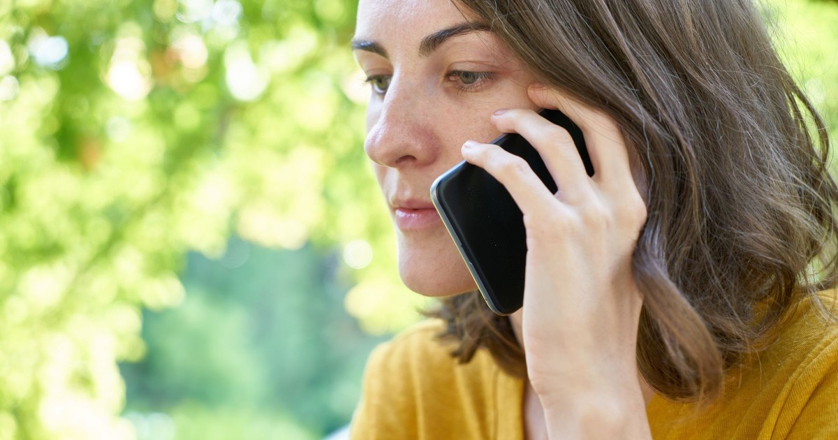 Side profile of a woman using a smartphone outdoors in a sunlit park, wearing a yellow top.