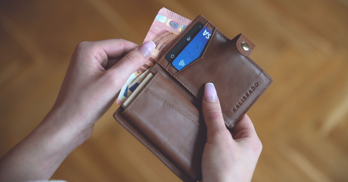 Hands holding a leather wallet with cash and a credit card on a wooden surface.