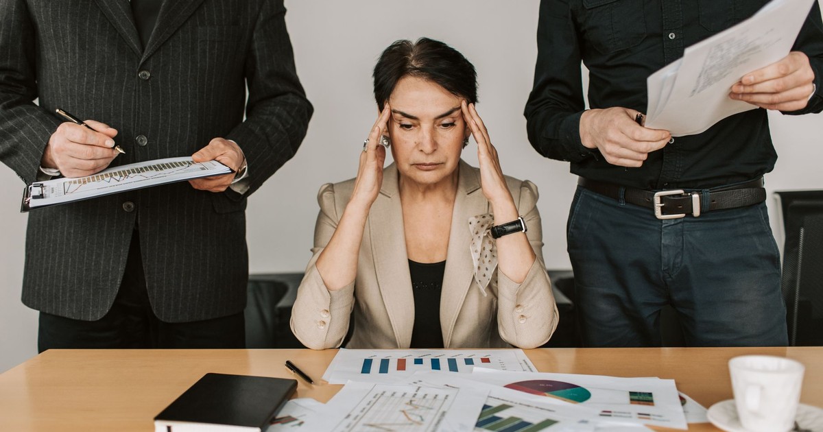A stressed businesswoman holding her head with colleagues holding documents in a corporate office setting.