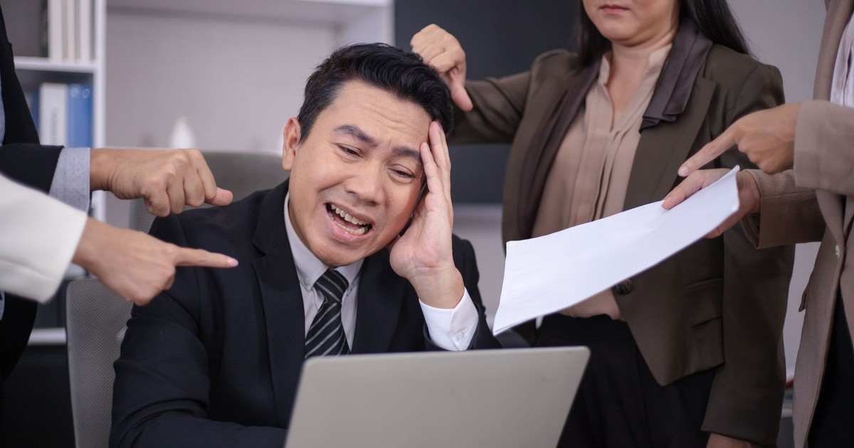 An Asian businessman looks stressed as colleagues point fingers and give him paperwork in a modern office.