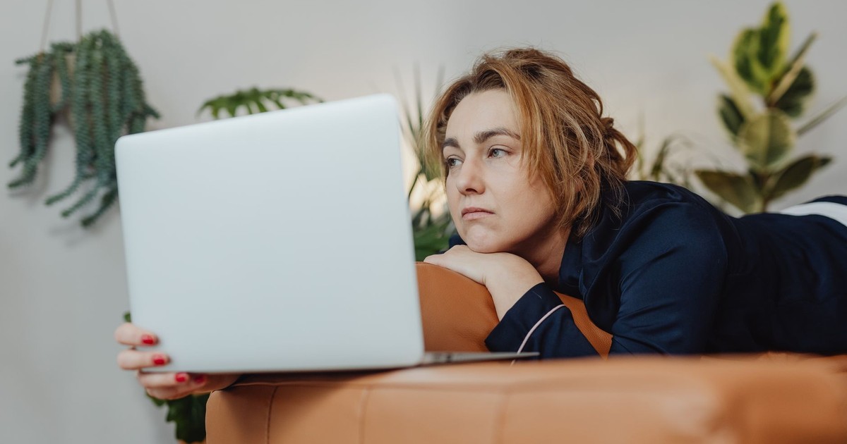 A woman lying on a couch indoors, looking upset while holding a laptop in a cozy home environment.