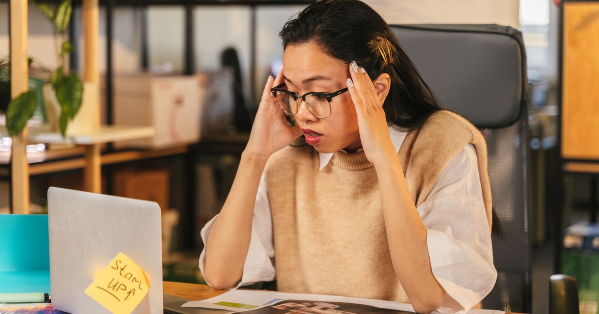 A woman with glasses shows stress while working at a laptop in an office environment.