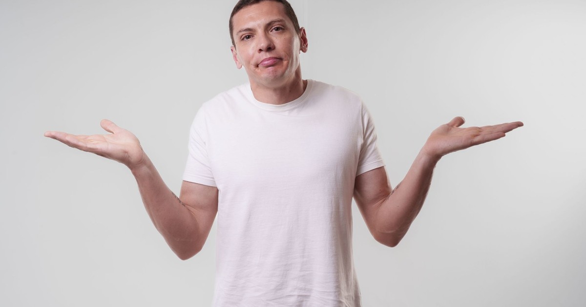 An adult man wearing a white t-shirt shrugs against a neutral background, conveying confusion or indifference.