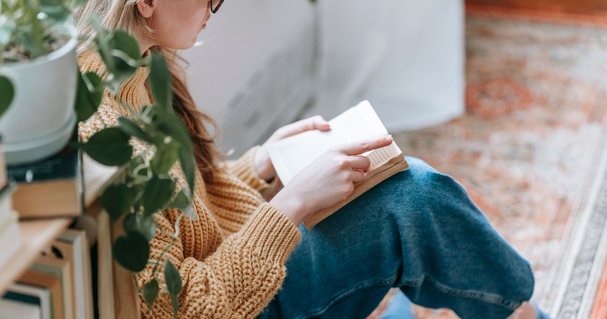 Side view of crop female student in casual clothes sitting on carpet near bookshelf with plant and reading book at home