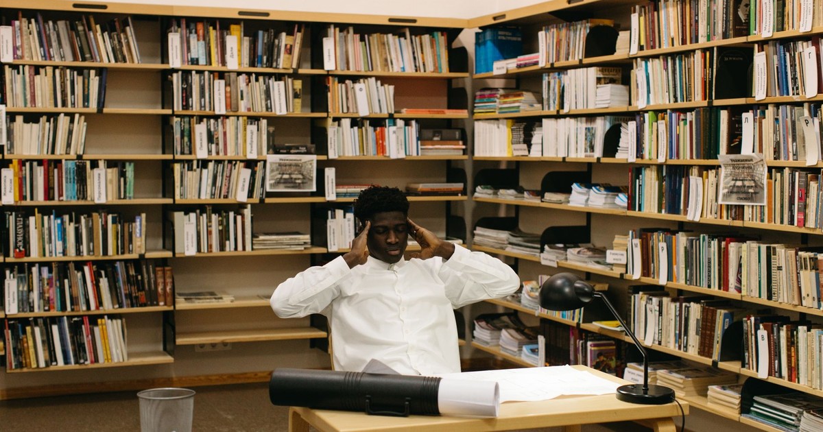 A student seated at a desk in a library, surrounded by bookshelves, concentrating with fingers on temples.