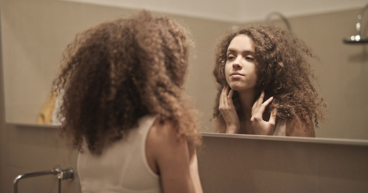 A woman with curly hair contemplates in front of a bathroom mirror, reflecting on her thoughts.