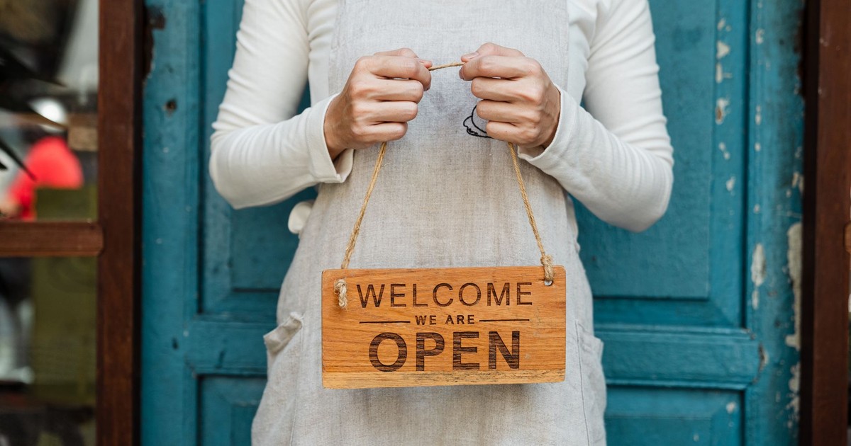 A person holds a "Welcome, we are open" sign in front of a rustic blue door.