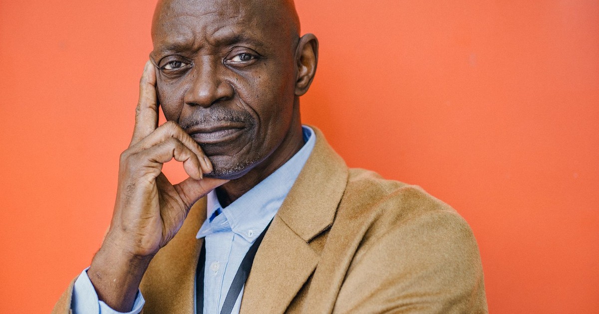 Confident middle aged bald African American male in elegant suit looking at camera with hand on cheek against orange background