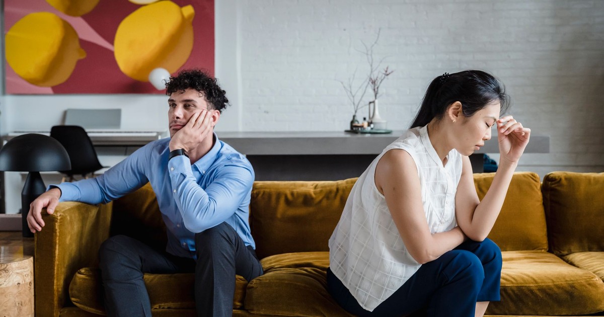 A couple sits apart on a sofa, looking unhappy and deep in thought, reflecting relationship conflict.