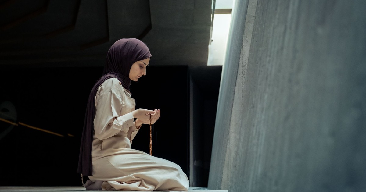 A woman in a hijab praying with rosary beads inside a modern, dimly lit building.