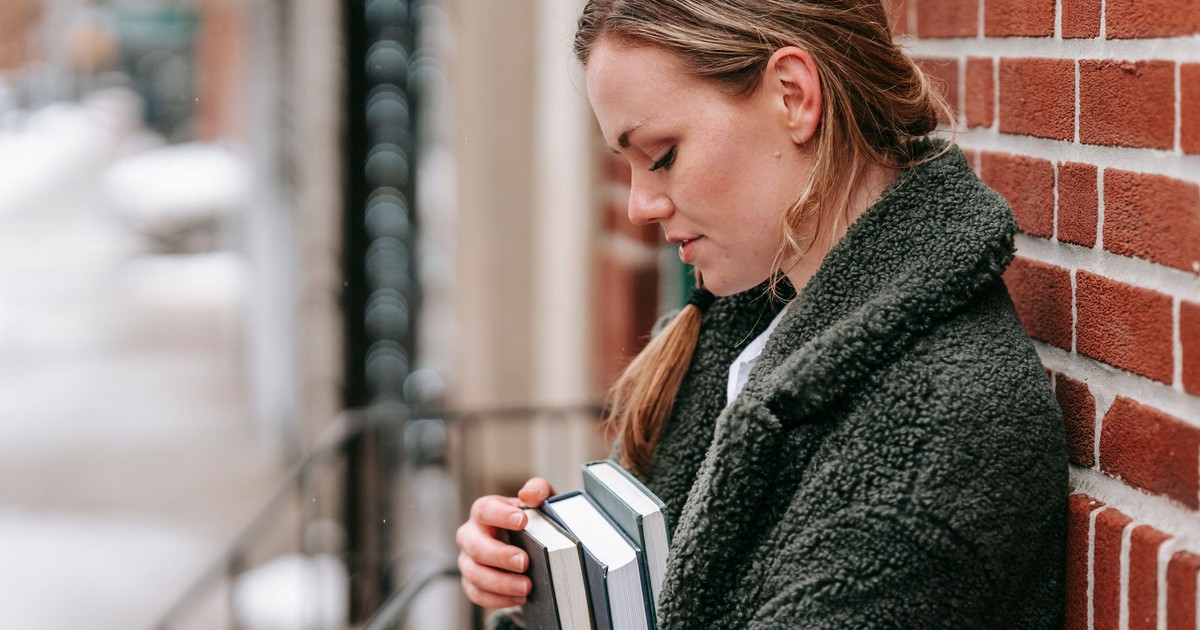 Side view of young female in outerwear with textbooks looking down against rough wall in town
