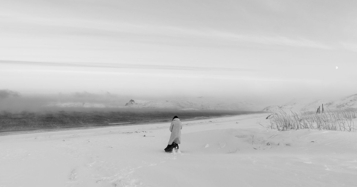 A lone person walks through a vast snowy terrain on a cold, foggy winter day.