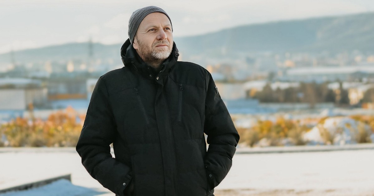A middle-aged man in a black jacket and beanie outdoors in a winter setting, looking into the distance.