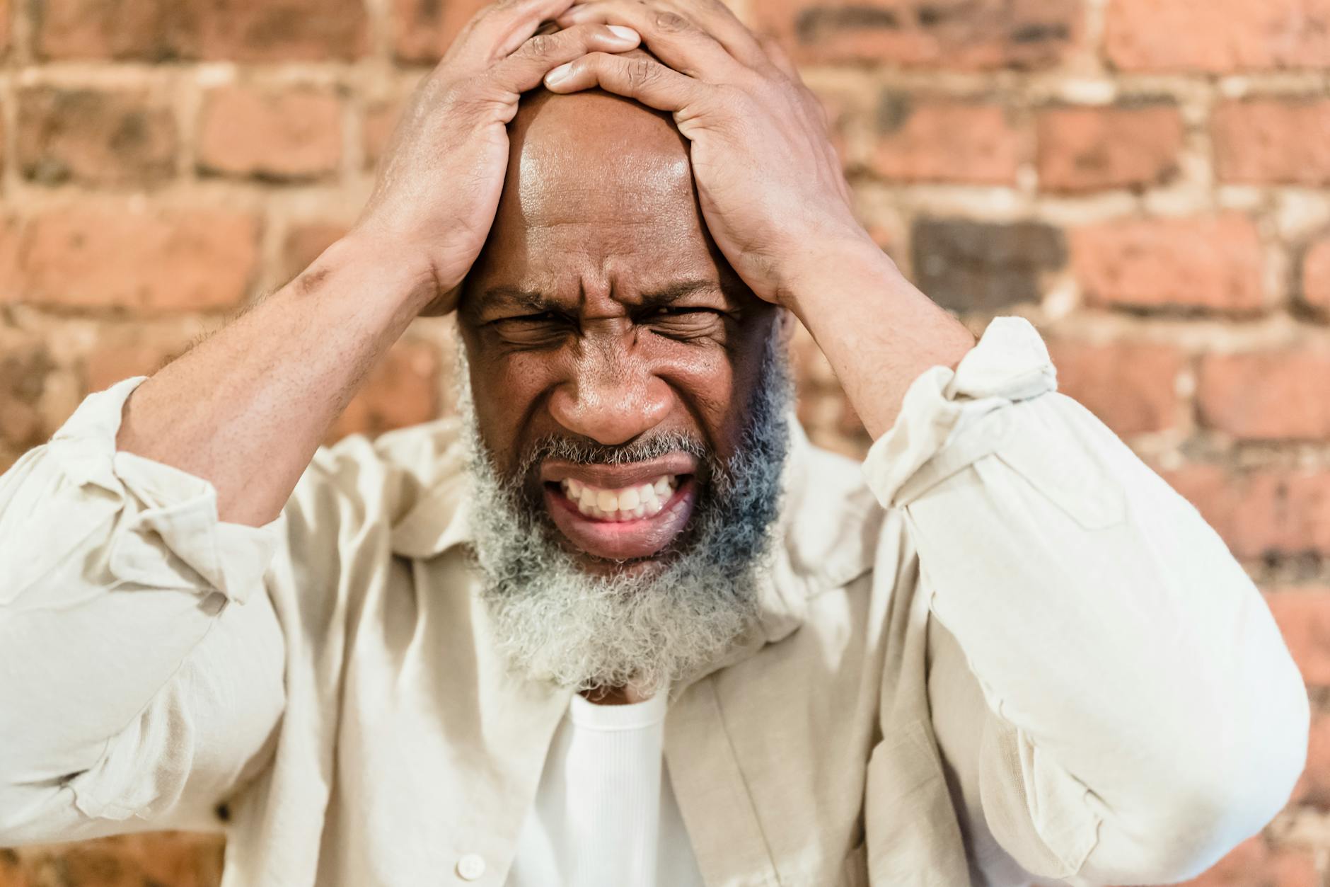 Close-up portrait of an adult man expressing pain and stress, holding head against a brick wall.