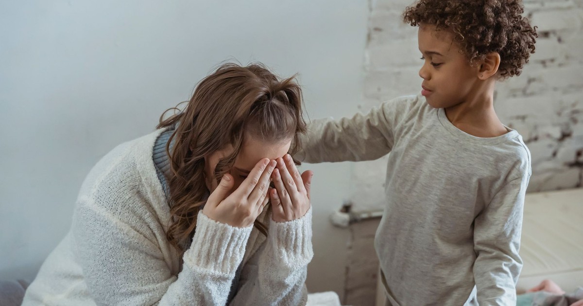 Caring African American son touching shoulder of upset faceless mother covering face while sitting in light room near wall at home
