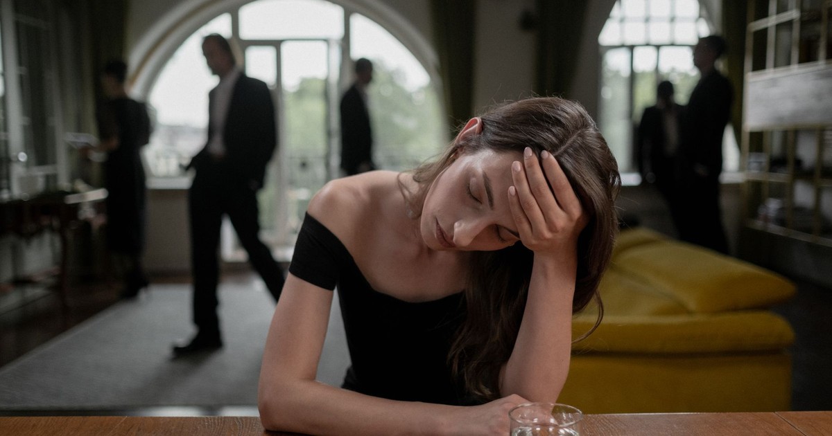 A stressed woman in a formal indoor setting, with a glass of water and blurred figures in the background.