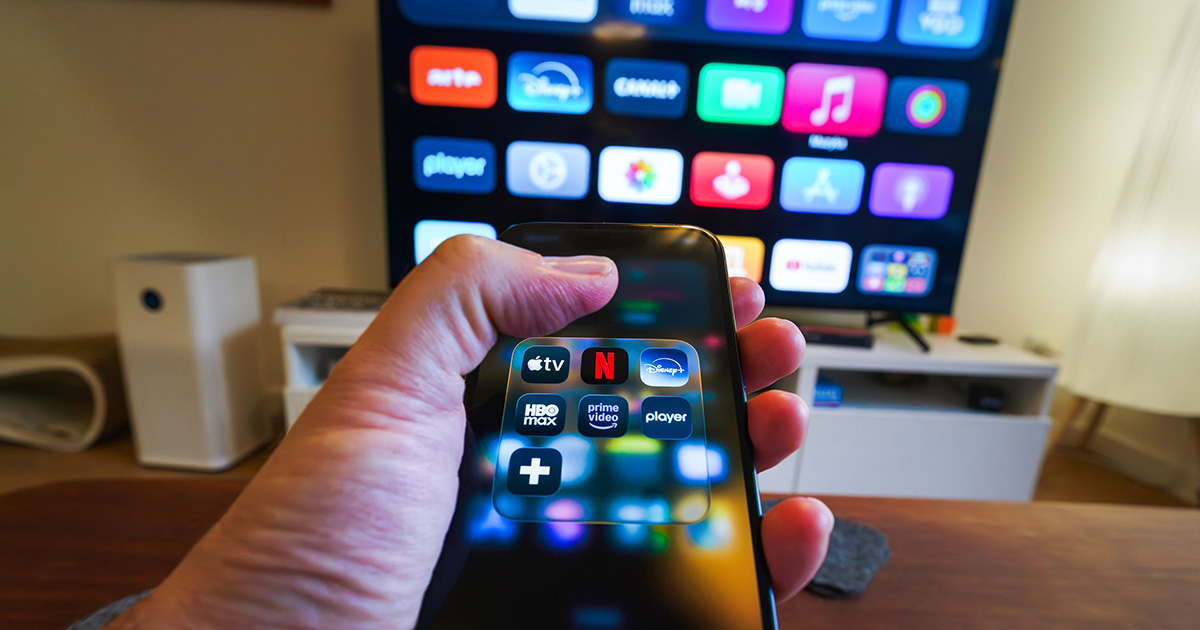 Close-up of a hand holding a phone displaying streaming apps in front of a TV with multiple app icons