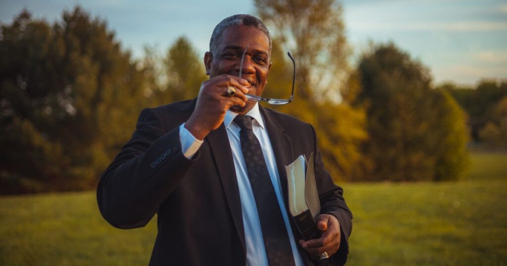 African American man in a suit smiling outside with eyeglasses and book