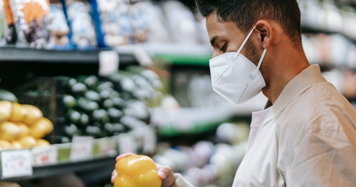 Side view of Indian male buyer in medical mask standing with yellow bell pepper in hand near stall with various vegetables on blurred background