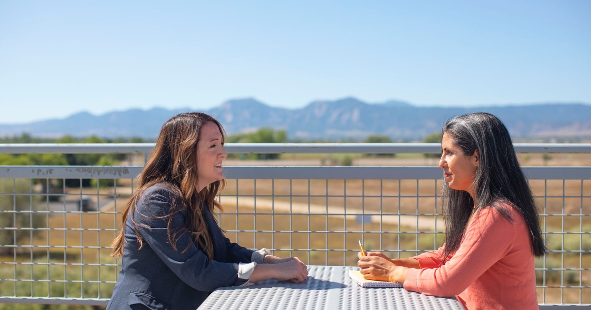 Two women discussing work at a table outdoors with a scenic mountain view.