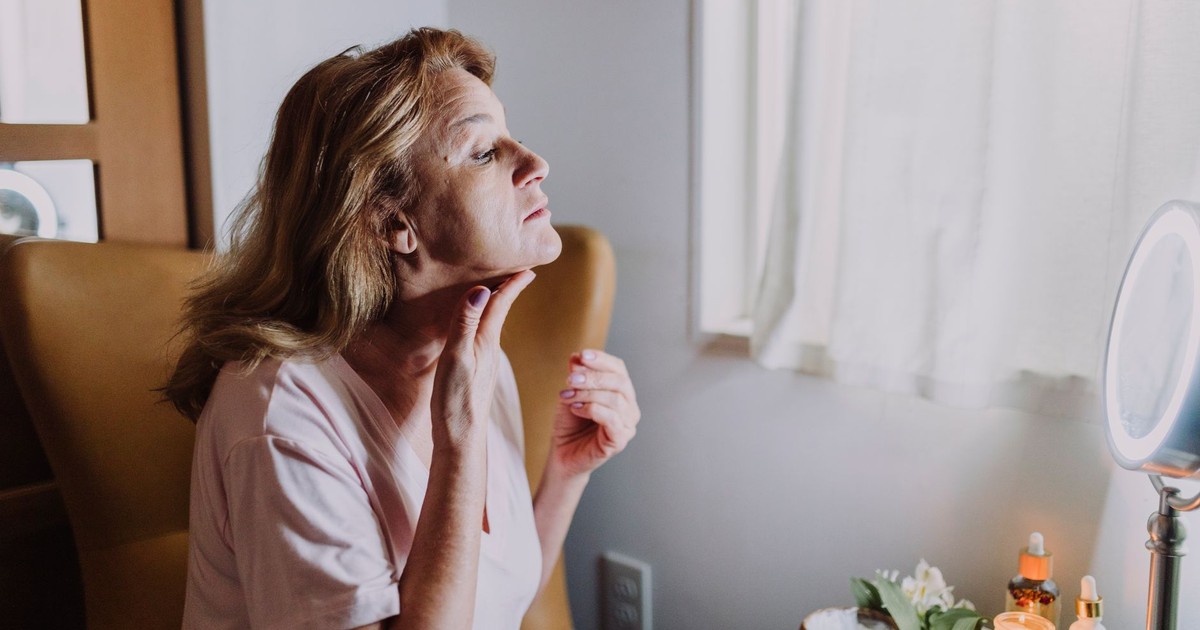 A mature woman examining her skin in a well-lit room with skincare products nearby.