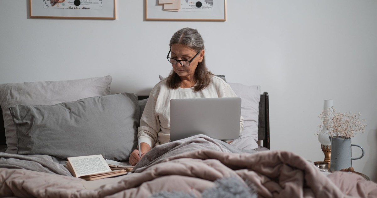 Elderly woman sitting on bed using laptop, writing notes. Comfortable home setting.
