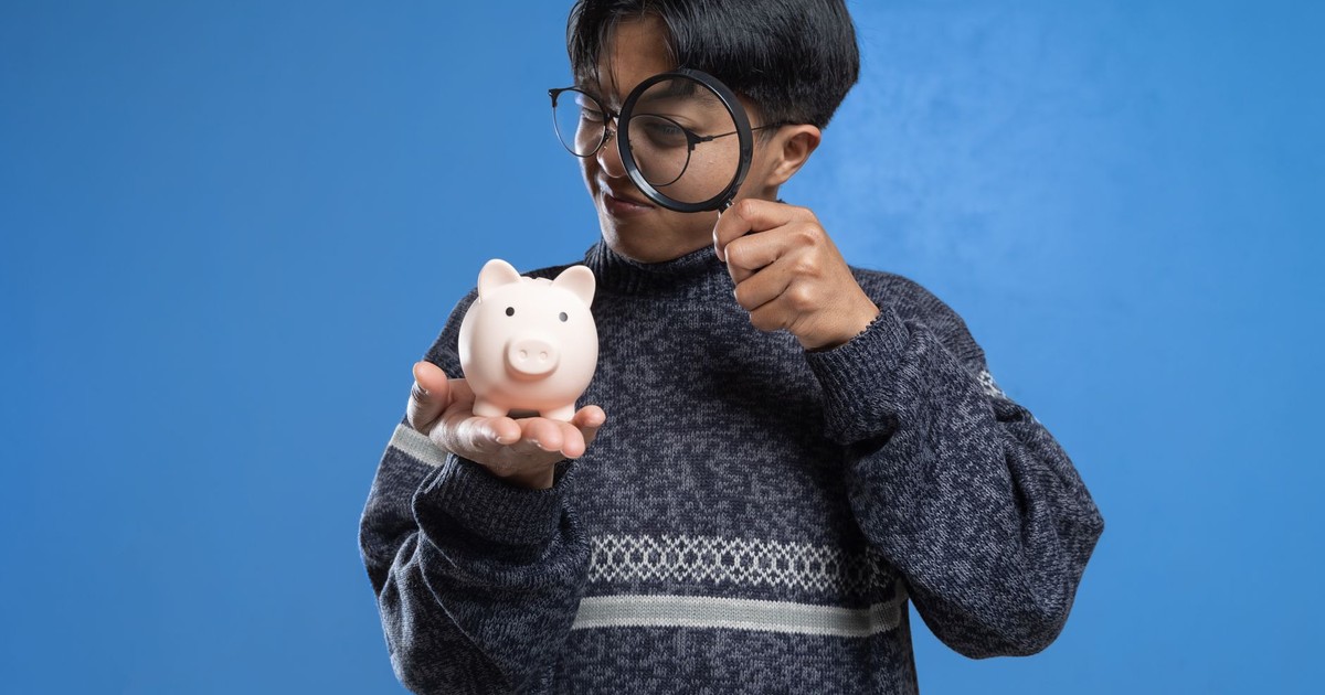 A person closely examines a piggy bank with a magnifying glass, symbolizing financial scrutiny.
