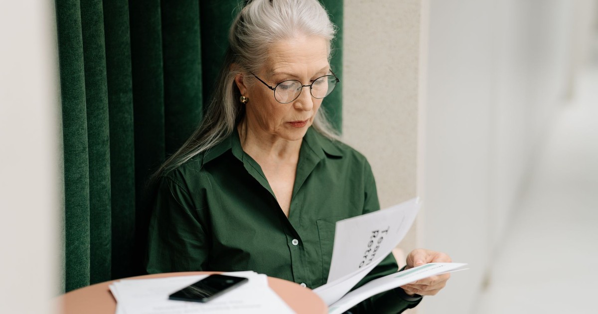 Elderly woman with gray hair reading papers at a small table indoors, focused and thoughtful.