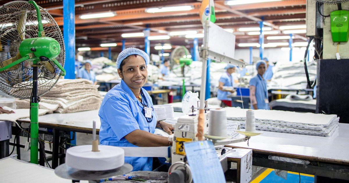 A female factory worker in India manages a sewing machine in a bustling textile factory.