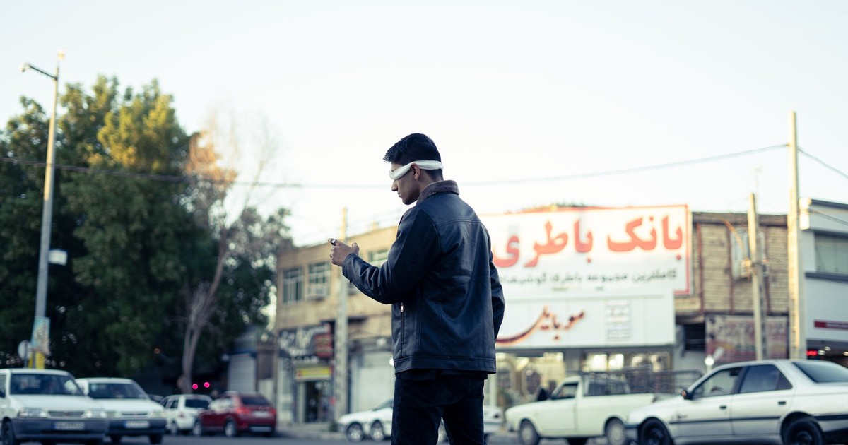 A man blindfolded walking across a city street in Jiroft, Iran, during the day.