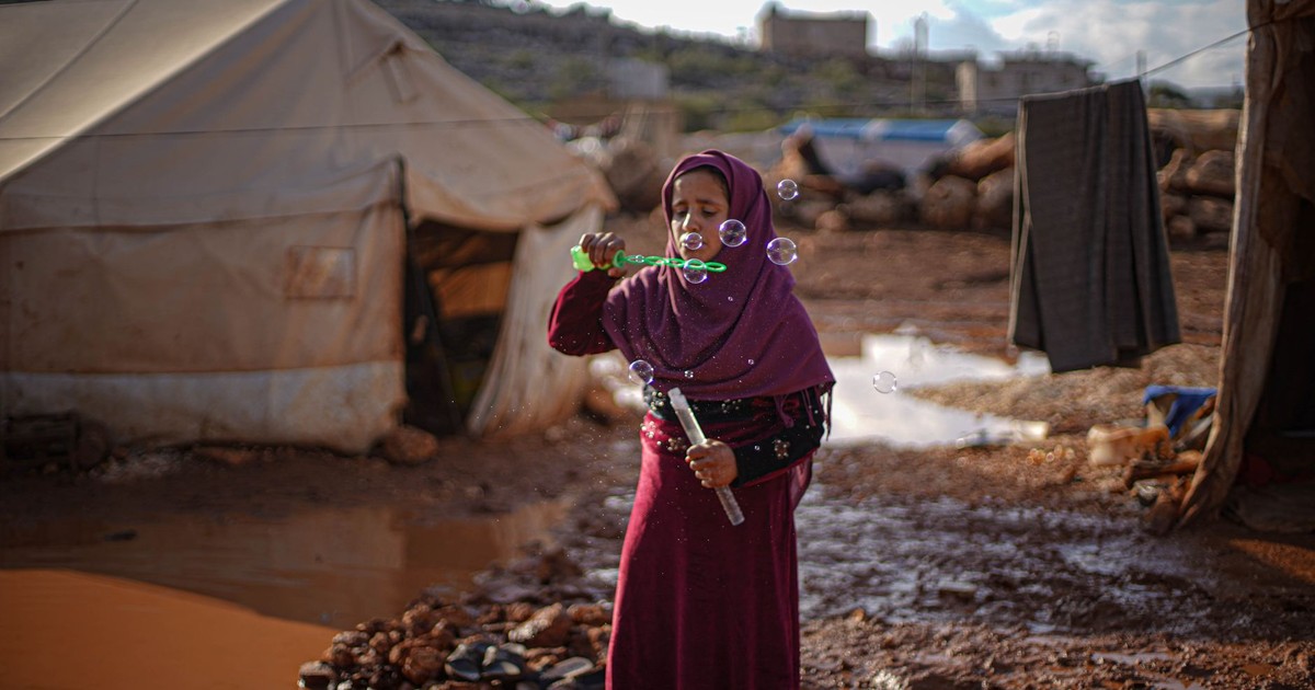 A girl in a Syrian refugee camp enjoys blowing bubbles amidst tents, capturing a moment of childhood joy.