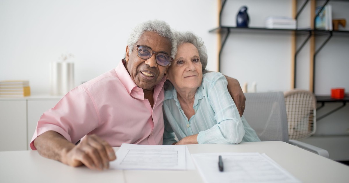 Elderly couple smiling and hugging while looking at documents on table indoors.