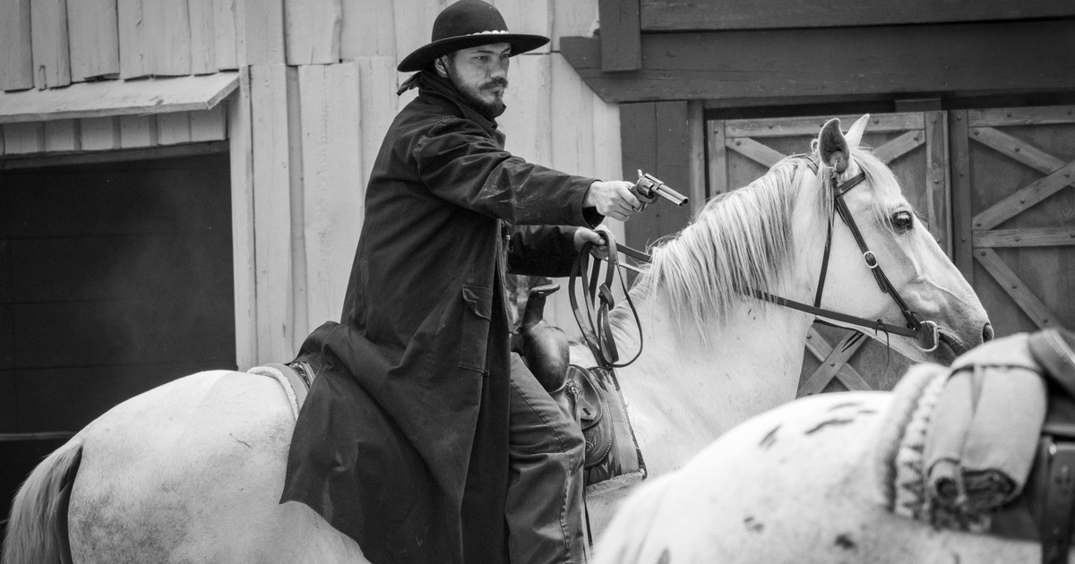 Black and white photo of a cowboy on horseback aiming a revolver in an old western setting.