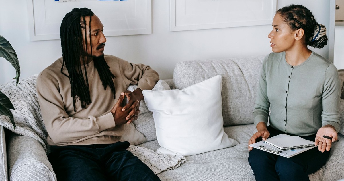 Distressed African American male in smart casual clothes sitting on comfy sofa and listening to professional female counselor in psychology center