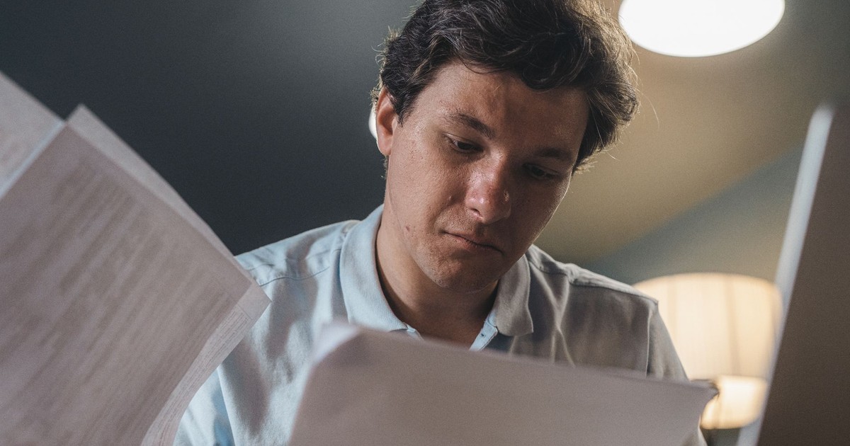 Serious man wearing a light blue shirt intently reading printed papers indoors under warm lighting.
